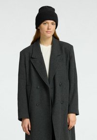 Sivi dvostruki kaput od teksturirane tkanine, s reverima i dva dugmeta, uparen s crnim pletenim beanie šeširom.