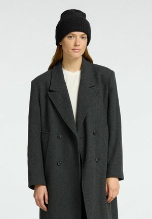 Cappotto grigio doppiopetto realizzato in tessuto strutturato, con rever a punta e due bottoni, abbinato a un berretto in maglia nera a coste.