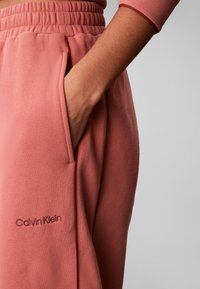 Χέρι στην τσέπη ροζ φόρμας Calvin Klein με ελαστική μέση και κέντημα λογότυπου στον αριστερό μηρό.