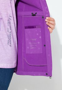 Henkilö, joka pitää avointa violettia takkia esillä, näyttäen sisä taskua, jossa on etiketti, joka ilmoittaa vedenpitävistä, tuulenpitävistä ja hengittävistä ominaisuuksista.