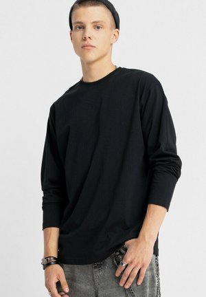 Młody mężczyzna w czarnej koszuli z długimi rękawami, czapce założonej tyłem, szarych dżinsach oraz z akcesoriami na nadgarstkach i palcach, stojący na tle białym.