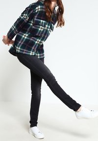 Koszula z flaneli w granatowo-zieloną kratę z dwoma kieszeniami na piersi, zestawiona z czarnymi obcisłymi dżinsami i białymi trampkami. Osoba jest w ruchu.