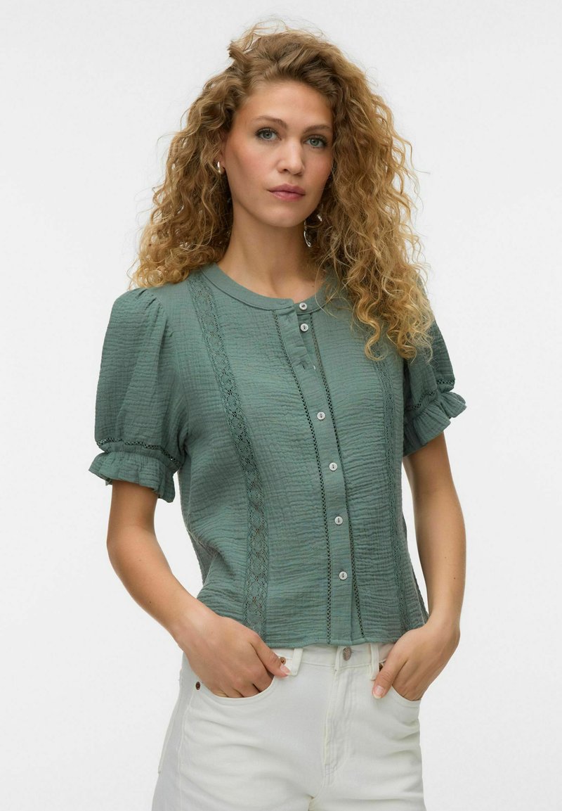 Femme aux cheveux blonds bouclés porte un chemisier vert texturé à manches courtes avec boutons et pantalon blanc, mains dans les poches, fond neutre.