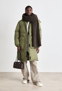 Olive green puffer coat with a zip front and pockets, paired with a dark brown textured scarf. Beige loose-fitting pants and white shoes.