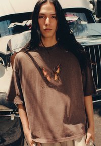 T-shirt oversized marrone con grafiche di farfalle in arancione e nero. Tessuto morbido, maniche corte e vestibilità comoda.