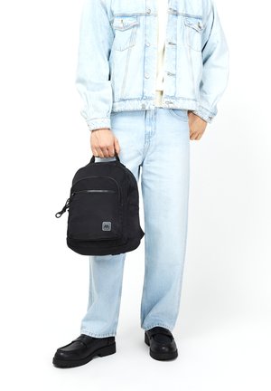 Mochila de tela negra con una textura suave, que cuenta con un bolsillo frontal con cremallera y un parche con el logo. El modelo viste una chaqueta de mezclilla y jeans claros.