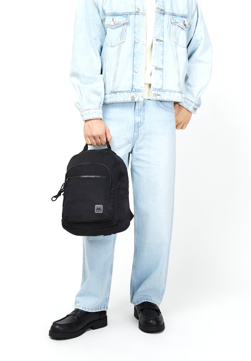 Černý textilní batoh s hladkým povrchem, vybavený přední kapsou na zip a nášivkou s logem. Model má na sobě džínovou bundu a světlé džíny.