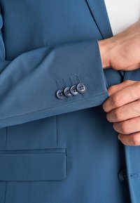 Persona che sistema il polsino di una giacca su misura blu con quattro bottoni abbinati e una tasca frontale visibile.