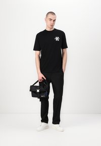 Μαύρο T-shirt με λογότυπο, μαύρο παντελόνι και μαύρη τσάντα με λουρί. Το μοντέλο φορά λευκά αθλητικά παπούτσια, στέκεται μπροστά από ένα απλό φόντο.