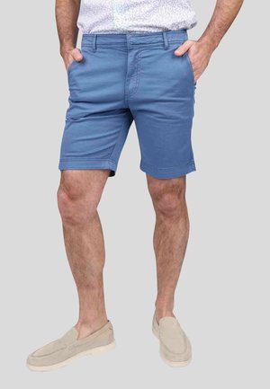 Shorts en coton bleu pour homme, coupe régulière, devant plat, poches latérales et passants de ceinture. Portés avec des chaussures à enfiler de couleur claire.