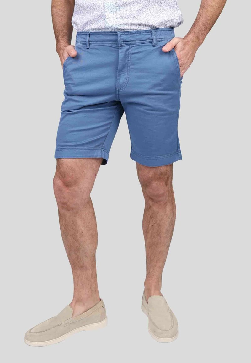 Shorts en coton bleu pour homme, coupe régulière, devant plat, poches latérales et passants de ceinture. Portés avec des chaussures à enfiler de couleur claire.