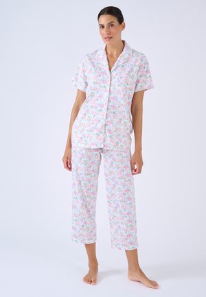Mujer con camiseta blanca de manga corta con botones y pantalones recortados con patrón floral rosa y azul, de pie descalza sobre fondo liso.