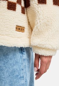 Giacca in pile morbido e color crema con motivo a scacchi marrone, dotata di un'etichetta rettangolare Roxy sulla manica sinistra in basso. Indossata con jeans blu chiaro.