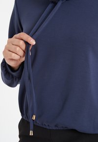 Marineblaues Langarmoberteil mit gerafftem Saum, das einen Kordelzug am Halsabschluss und goldfarbene Akzente an den Kordelenden aufweist.