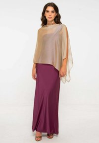 Femme portant un châle en beige transparent sur une robe sans manches mauve et des sandales dorées, se tenant devant un fond blanc uni.