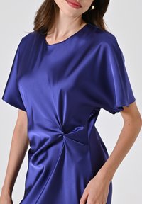 Abito di raso di un profondo colore viola con maniche corte e un design intrecciato in vita, caratterizzato da una trama liscia e un colletto rotondo.