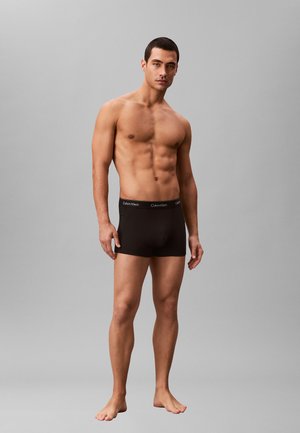 Svarta bomullsboxershorts med ett elastiskt midjeband med en logotyp. Modellen står barfota och visar upp en åtsittande design och en slät textur.