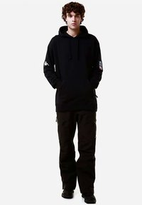 Felpa nera con cappuccio, tasca frontale e coulisse, con un logo bianco sulla manica sinistra. Abbinata a pantaloni cargo neri.