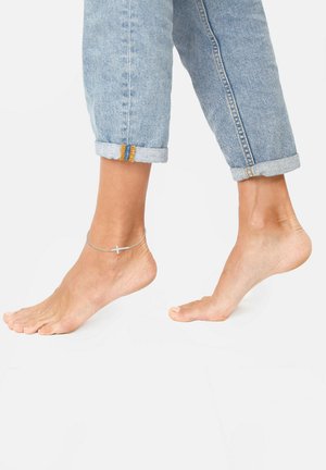Bracciale alla caviglia in argento con un piccolo dettaglio a croce, indossato su un piede nudo. Jeans blu arrotolati con cuciture a contrasto visibili vicino al fondo.