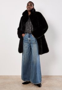 Schwarzer Kunstfellmantel mit großem Kragen, kombiniert mit einem silbernen Paillettenoberteil und weiteren blauen Denim-Jeans. Schwarze Schuhe vervollständigen das Outfit.