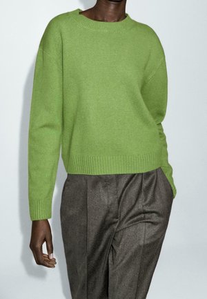 Personne portant un pull en maille vert clair à manches longues et un pantalon gris plissé taille haute, une main dans la poche.