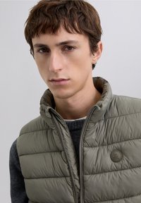 Lysegrøn quiltet dunvest med høj krave, med lynlås foran og rundt logoemblem. Båret over en mørkegrå trøje.