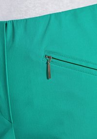 Tissu bleu sarcelle avec une texture lisse, présentant une poche zippée avec quincaillerie argentée et un design de bandeau plat en élastique.