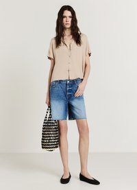 Chemise beige à manches courtes avec boutons, short en jean bleu clair, chaussures plates noires et sac fourre-tout noir et blanc à motifs.