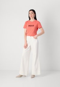 Sieviete stāv minimālisma stilā, ģērbta korallkrāsas T-kreklā ar uzrakstu "BOSS", baltās brīvās biksēs un bēšās kurpēs ar augstiem papēžiem.