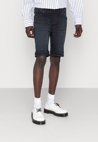 Mörka jeanstorts med en figurnära design och uppvikta ärmar, kombinerat med vita skor och strumpor, mot en neutral bakgrund.