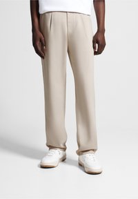 Beige trousers with front pleats, a straight fit, and a smooth texture, paired with white sneakers featuring a gum sole.