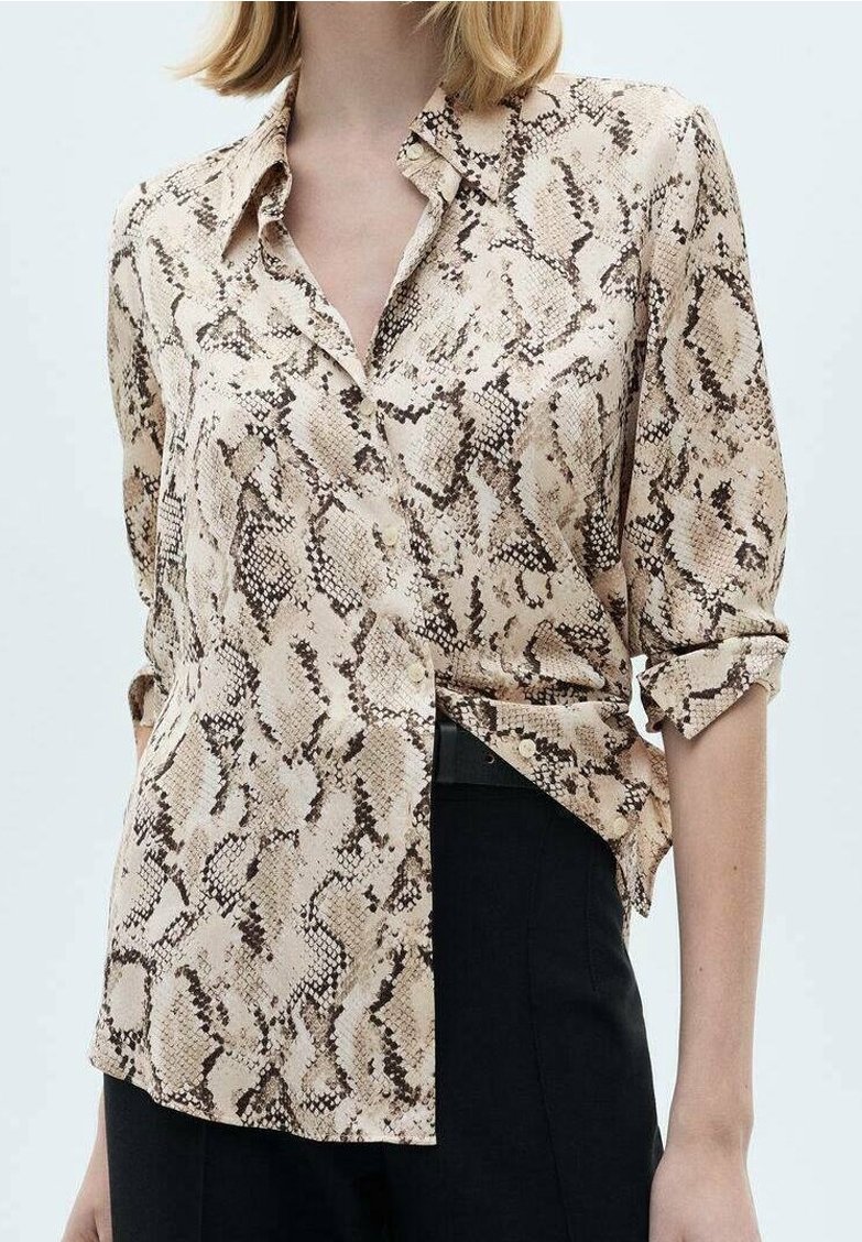 Femme portant une chemise boutonnée à manches retroussées avec un imprimé serpent beige et marron, rentrée dans un pantalon noir, sur un fond uni.