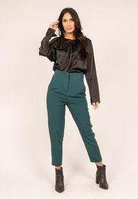 Haut noir en satin à manches longues avec un col froncé, associé à un pantalon vert tapered taille haute et des bottines noires.