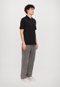 Svart polo skjorte med glidelås krage, kombinert med grå denim bukser. Modellen har på seg hvite sneakers, synlige sømmer og en avslappet passform.