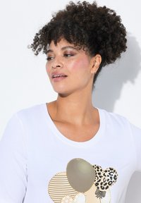 La camiseta blanca presenta un diseño impreso con diversas formas circulares en dorado, negro y estampado animal, adornado con acentos brillantes.
