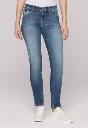 Person wearing slim-fit blue jeans and white sneakers against a plain light background, showing the lower body front view.