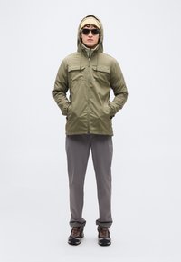 Πράσινο μπουφάν ελιάς με κουκούλα, εμπρόσθιες τσέπες και κλείσιμο με φερμουάρ, συνδυασμένο με ανοιχτό γκρι παντελόνι και καφέ παπούτσια πεζοπορίας. Φορεμένα μαύρα γυαλιά ηλίου.