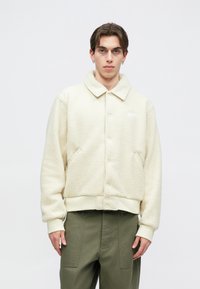 Μπουφάν από γαλότσες σε κρεμ χρώμα με δομημένο γιακά, κουμπιά μπροστά και πλαϊνές τσέπες. Οι μανσέτες και το τελείωμα είναι ριμπ. Διακριτικό λογότυπο στο στήθος.