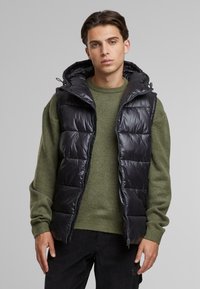Svart, puffy, ermeløs vest med hette, med quiltet design og glidelåsforclosing, båret over en grønn strikkegenser.