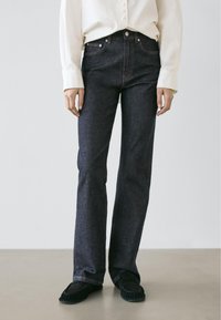 Jeans in denim scuro con vestibilità dritta, caratterizzati da un classico design a cinque tasche e cuciture sobrie, abbinati a una camicia leggera con bottoni.