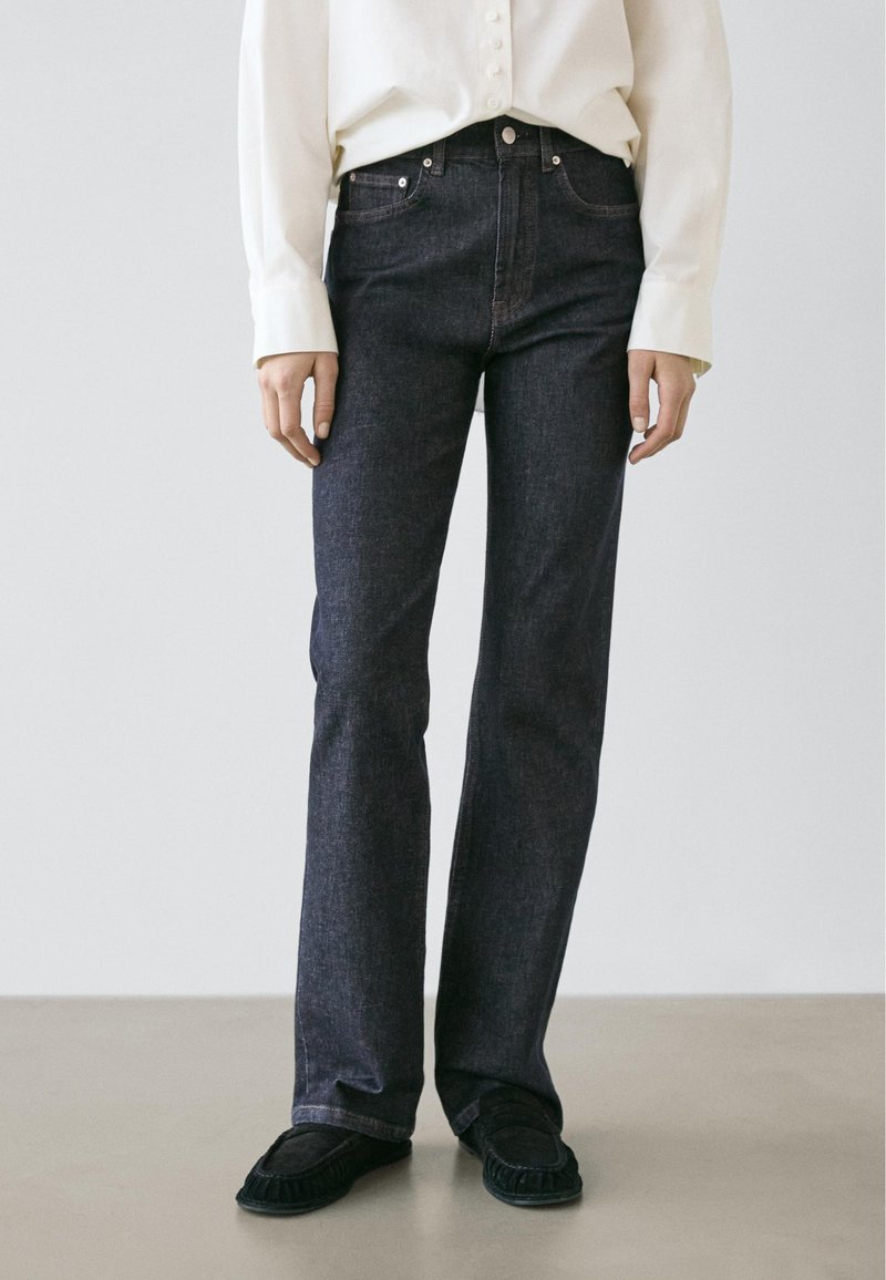 Jeans in denim scuro con vestibilità dritta, caratterizzati da un classico design a cinque tasche e cuciture sobrie, abbinati a una camicia leggera con bottoni.