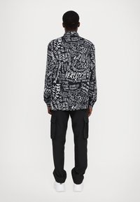 Schwarzes Langarmshirt mit weißen grafischen Textmustern, kombiniert mit schwarzen Cargo-Hosen und weißen Sneakern. Lässiges, modernes Design.