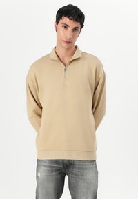 Felpa beige con zip e colletto alto, dalla texture morbida e orlo a coste. Presenta un piccolo logo sopra il petto e spalle scese.
