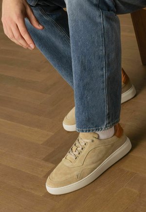 Beige suède sneakers met getextureerde tan leren accenten en witte rubberen zolen, gecombineerd met blauwe denim jeans, op een houten vloer.