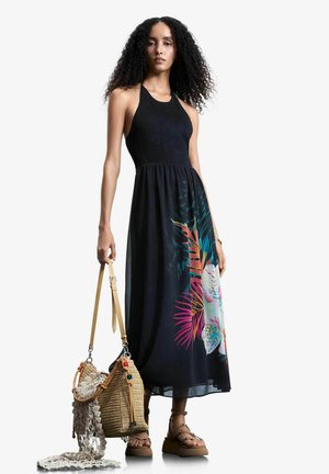 Mujer con cabello rizado vistiendo un vestido maxi negro sin mangas con estampado floral colorido, sosteniendo una bolsa tejida con tela de red y usando sandalias.