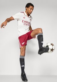 Camisola branca da Adidas com detalhes em bordô, texto "Emirates Fly Better", calções vermelhos, meias pretas com riscas brancas, e chuteiras pretas, a chutar uma bola de futebol.