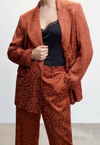 Un costume en velours côtelé orange brûlée avec un motif texturé à pois, composé d'une veste taillée et d'un pantalon assorti, superposé sur un top en dentelle noire.