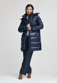 Granatowa kurtka puchowa z wysokim kołnierzem, błyszczącą pikowaną teksturą, zamkiem błyskawicznym i kieszeniami bocznymi, zestawiona z ciemnymi spodniami i brązowymi botkami za kostkę.