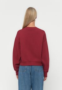 Sweatshirt bordeaux court à manches longues, avec col côtelé et ourlets à poignet, associé à un jean bleu taille haute, vu de l'arrière.