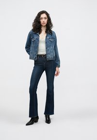 Jeans jakna v srednje modri barvi z gumbi spredaj, prsni žepi in dolgimi rokavi, kombinirana s temno modrimi razširjenimi hlačami in črnimi škornji z visokimi petami.